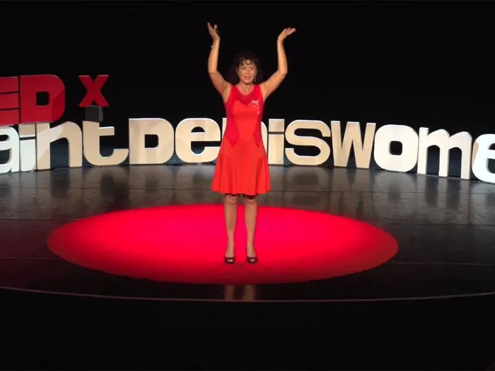 Miliana CORNESANU, conférencière, se tient sur la scène de TEDx en robe rouge, levant les bras en signe de victoire. Elle partage son expérience sur l'importance de suivre son cœur et la lumière intérieure pour transformer sa vie personnelle et professionnelle, mettant en lumière comment son coaching avec Kris Saint Ange l'a aidée à surmonter des défis et à trouver un équilibre émotionnel et professionnel.