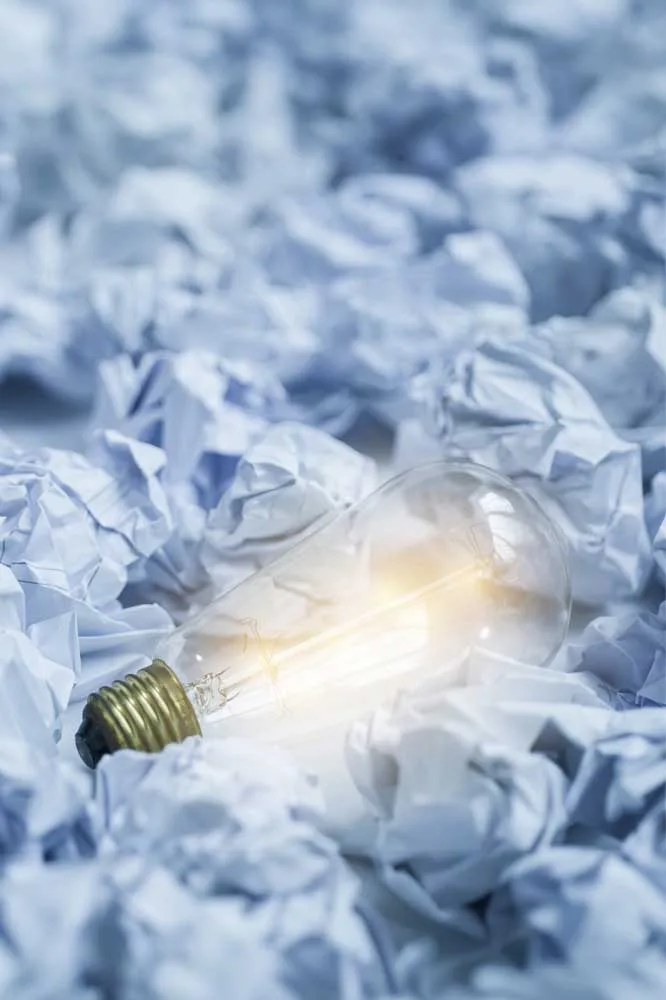 Image d'une ampoule illuminée au milieu de papiers froissés, symbolisant la catégorie 'Coaching' sur le blog de Kris Saint Ange. Cette lumière représente les idées et les solutions innovantes qui émergent dans le processus de coaching pour surmonter les défis et catalyser le développement personnel et professionnel.