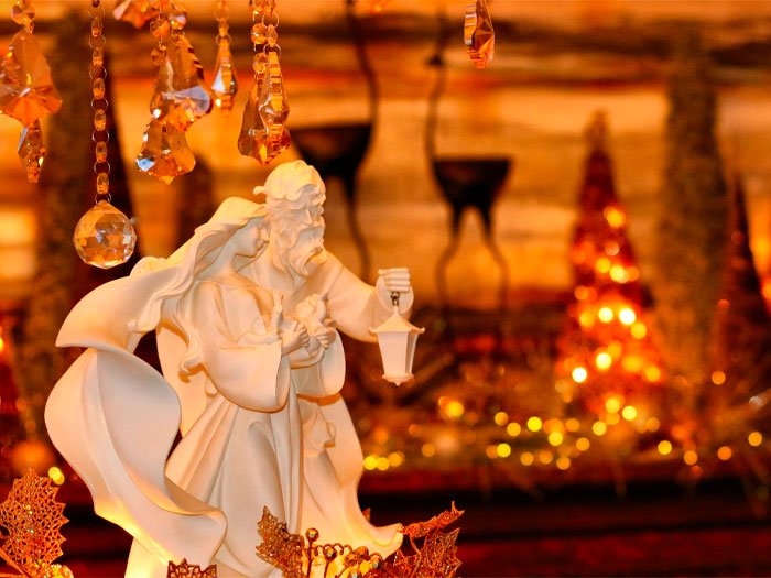 Statue en céramique de Joseph et Marie, portant une lanterne, placée devant un décor de Noël étincelant avec des lumières dorées et des ornements, symbolisant la lumière et l'espoir de la saison.