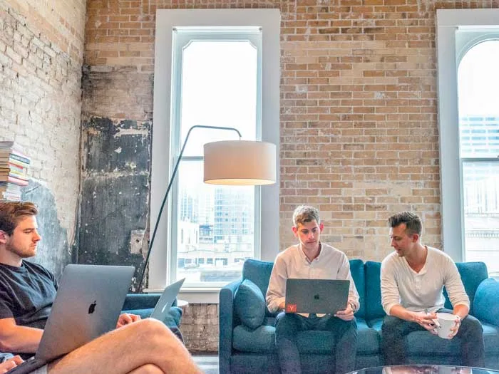 Trois jeunes hommes travaillent dans un espace de bureau moderne avec des murs en brique apparente, utilisant des ordinateurs portables. Ils représentent l'intégration de l'intuition et des stratégies collaboratives dans le processus de recrutement, symbolisant une approche innovante de la gestion des ressources humaines.